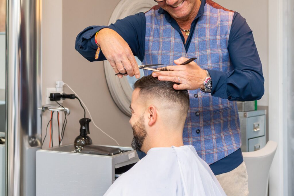 Un parrucchiere con un gilet a quadri blu taglia i capelli a un cliente usando forbici e pettine in un salone. Il cliente ha i capelli corti, la barba e indossa una mantella da taglio bianca.