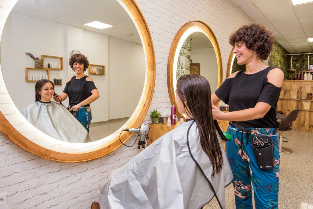 Una parrucchiera sorridente con capelli corti ricci spazzola i capelli lunghi e umidi di una cliente davanti a un grande specchio rotondo illuminato. La scena si riflette nello specchio, mostrando l'interno del salone.