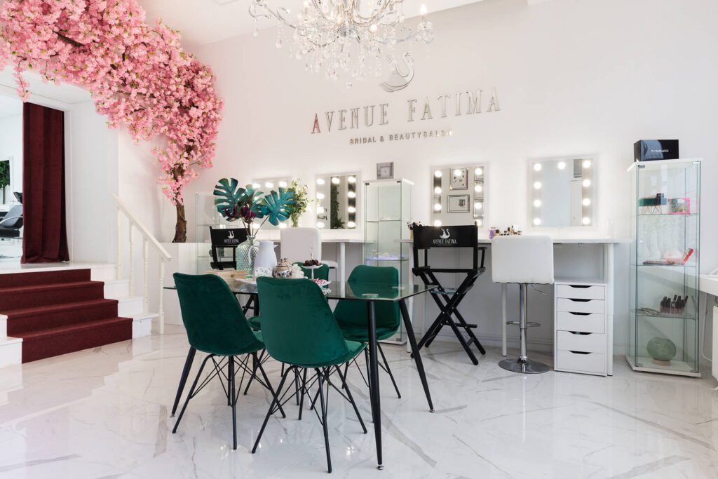 Un salone di bellezza e acconciatura sposa luminoso ed elegante chiamato "Avenue Fatima". Lo spazio presenta diverse postazioni trucco con specchi illuminati, sedie in velluto verde smeraldo attorno a un tavolo di vetro e un albero di ciliegio rosa decorativo.