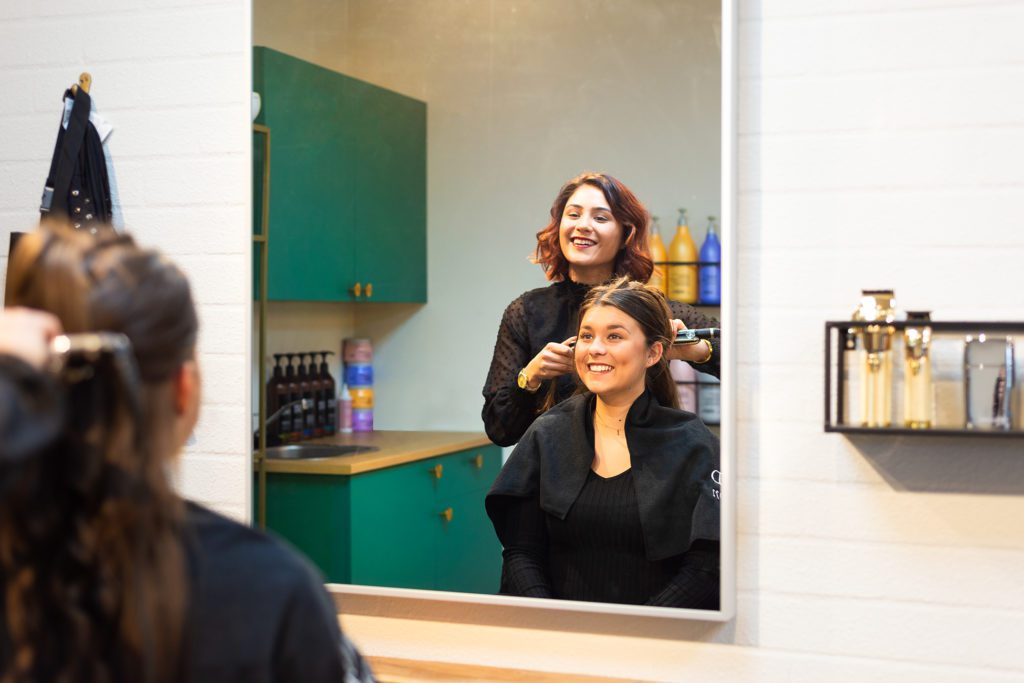 Una parrucchiera sorridente con capelli ramati acconcia i capelli di una cliente sorridente con una piastra, vista attraverso il riflesso di un grande specchio rettangolare in un salone moderno con mobili verde smeraldo.