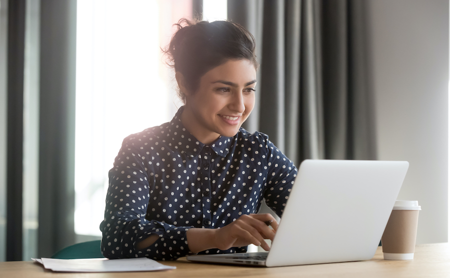 Una donna sorridente con i capelli raccolti in uno chignon, che indossa una camicia blu scuro a pois, siede a una scrivania di legno mentre scrive su un laptop d'argento accanto a una tazza di caffè da asporto.