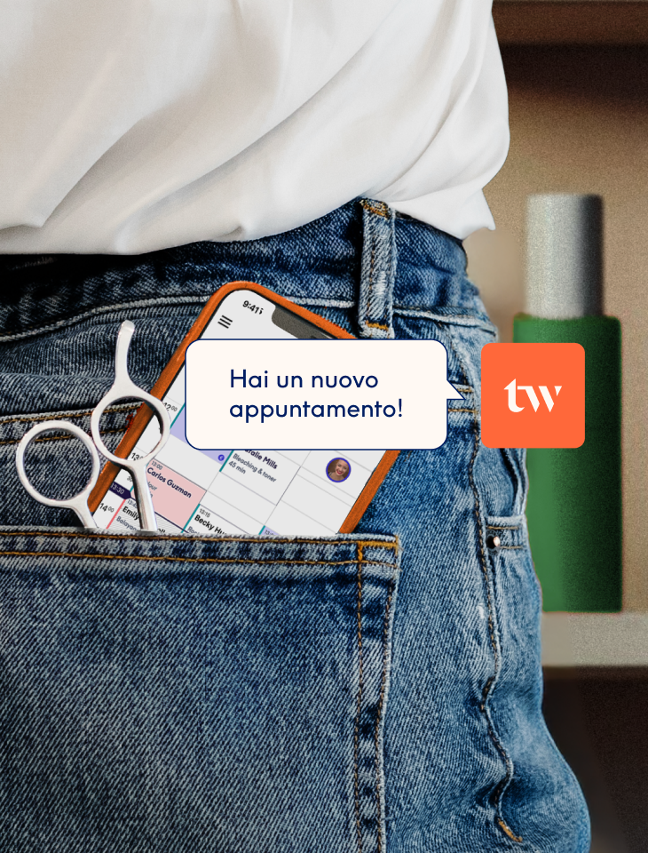 Uno smartphone e un paio di forbici da parrucchiere infilati nella tasca posteriore dei jeans, con lo schermo che mostra l'agenda delle prenotazioni dell'app Treatwell e la notifica "hai un nuovo appuntamento!".