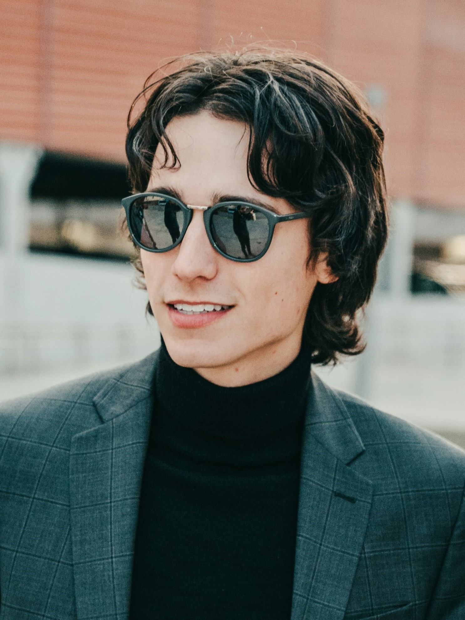 Un giovane uomo con un mullet retrò, giacca grigia e occhiali da sole, che sorride in piedi all'aperto.