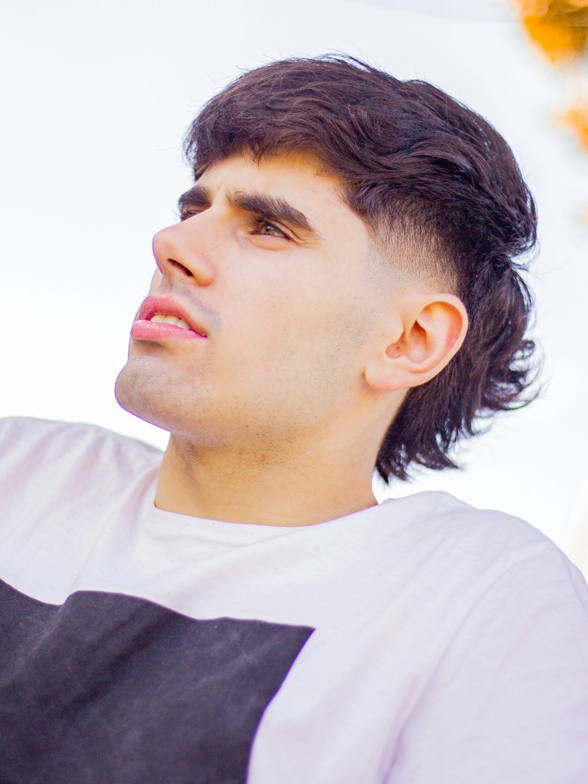 Un giovane uomo con una t-shirt bianca e un fade mullet definito, seduto vicino a una strada in una giornata di sole.