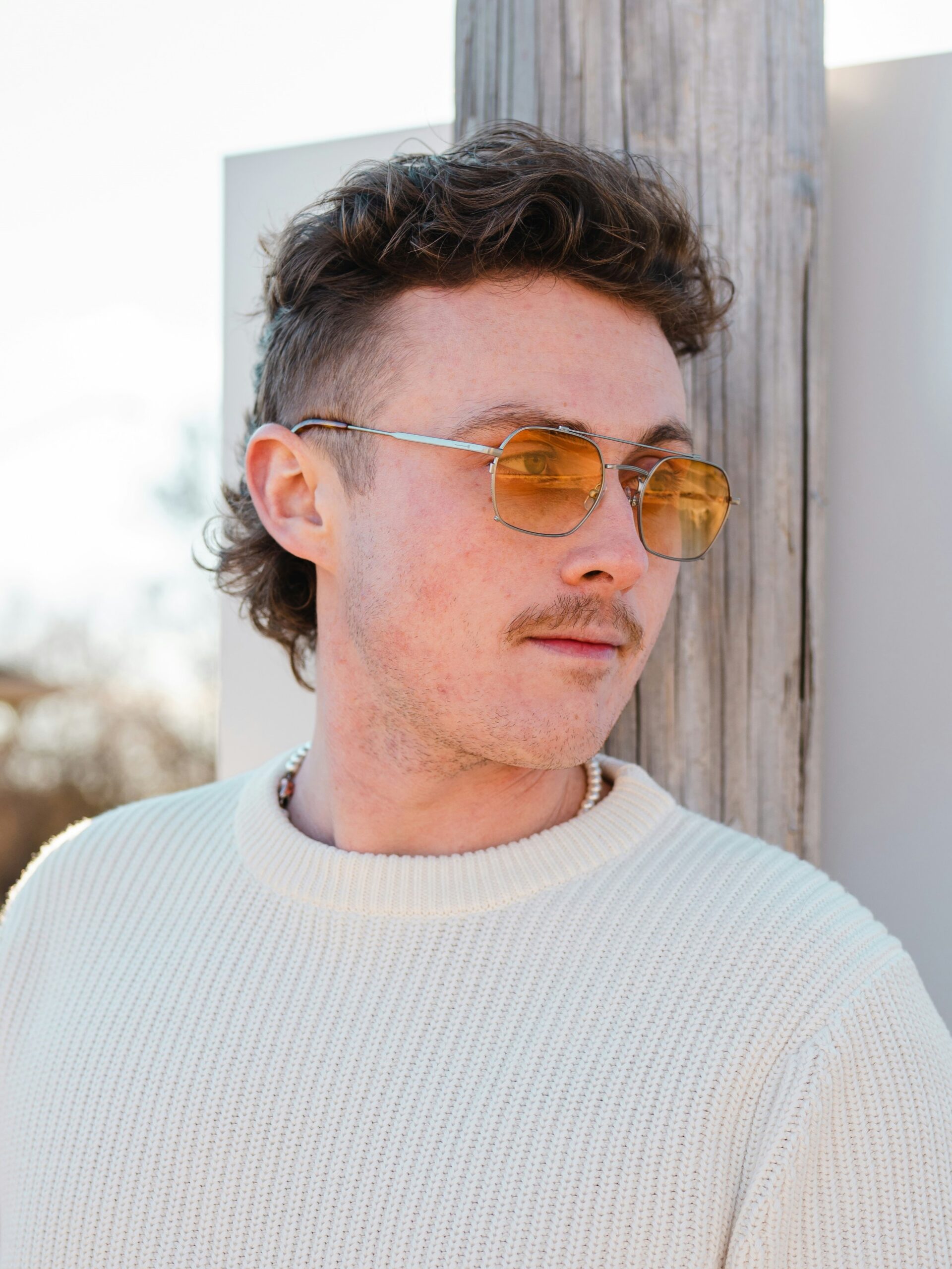 Un giovane uomo con un mullet riccio appoggiato a un muro all'aperto, in una giornata di sole.