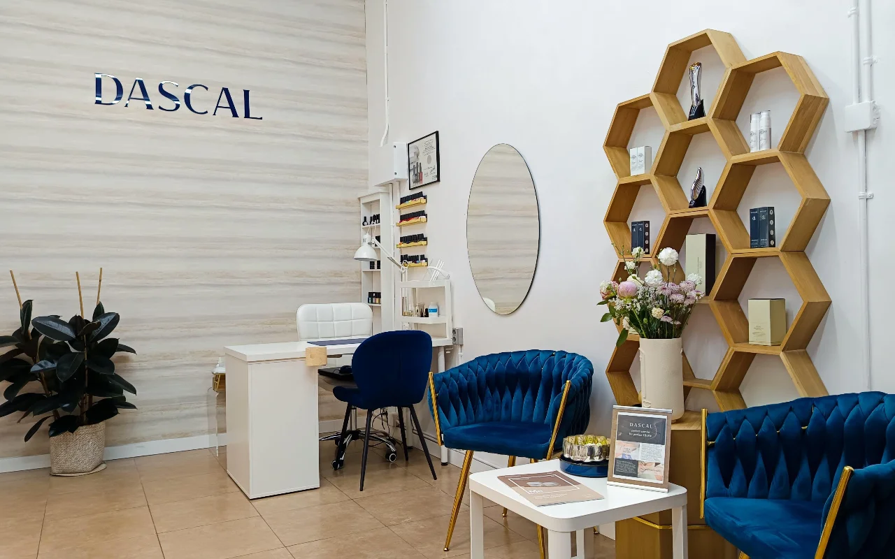 Un elegante interno di salone di bellezza con poltrone in velluto blu scuro e un originale scaffale esagonale in legno a parete. Un tavolo da manicure bianco si trova nell'angolo, addossato a una parete chiara dalla texture materica con la scritta "DASCAL" in lettere blu.