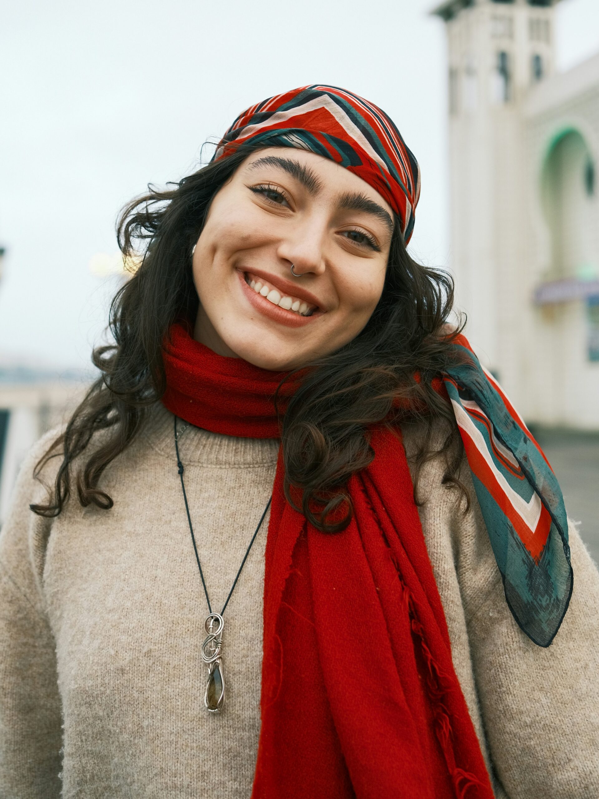 Donna sorridente in piedi all’aperto su un molo, con capelli scuri lunghi, un foulard a righe rosso, bianco e verde sulla testa e una sciarpa rossa al collo