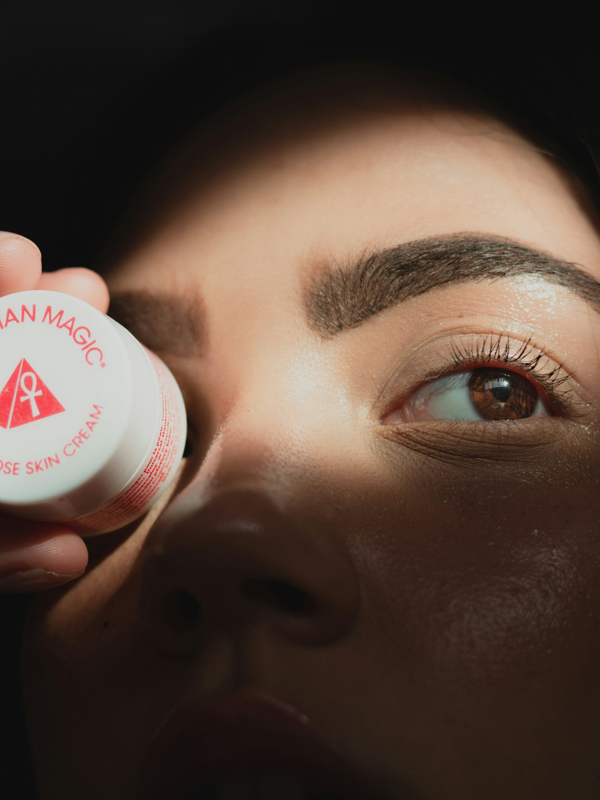 Primo piano del viso di una donna con microblading sulle sopracciglia che tiene un piccolo vasetto di crema davanti a un occhio
