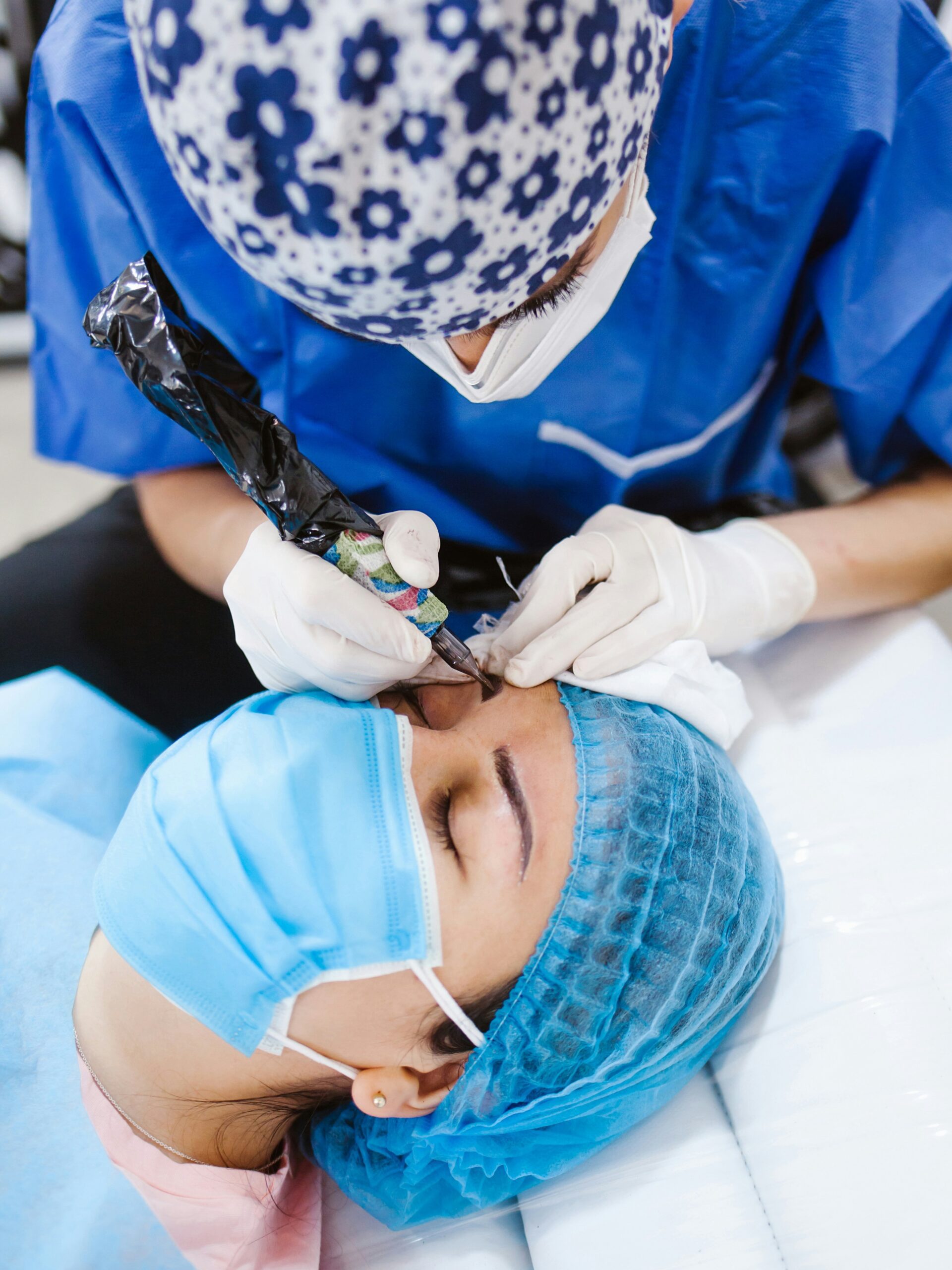 Primo piano di una donna durante un trattamento di microblading eseguito da un'operatrice; è distesa su un lettino e indossa una mascherina