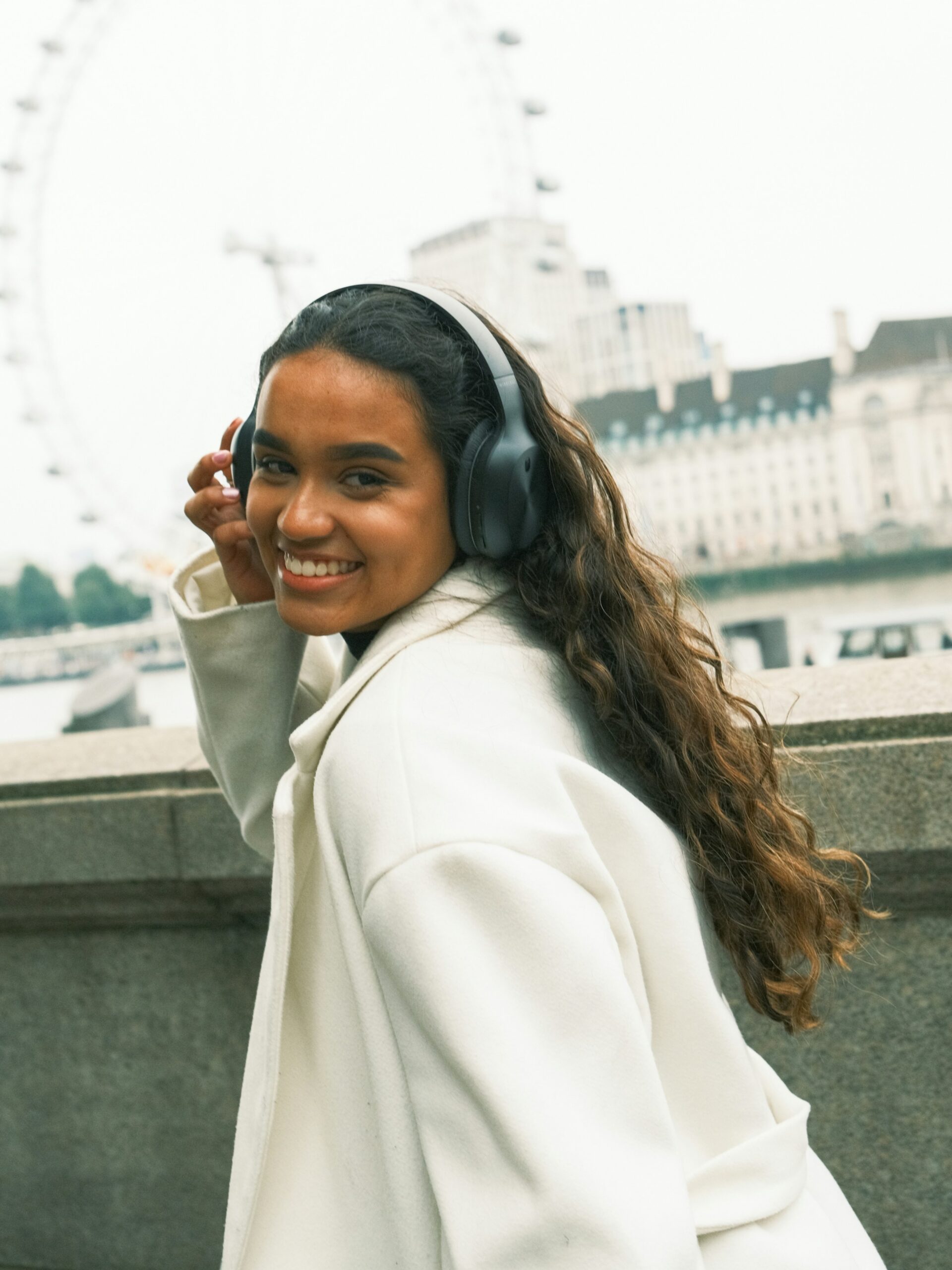 Una donna sorridente con microblading sulle sopracciglia, cuffie nere over-ear e cappotto bianco, in una città italiana