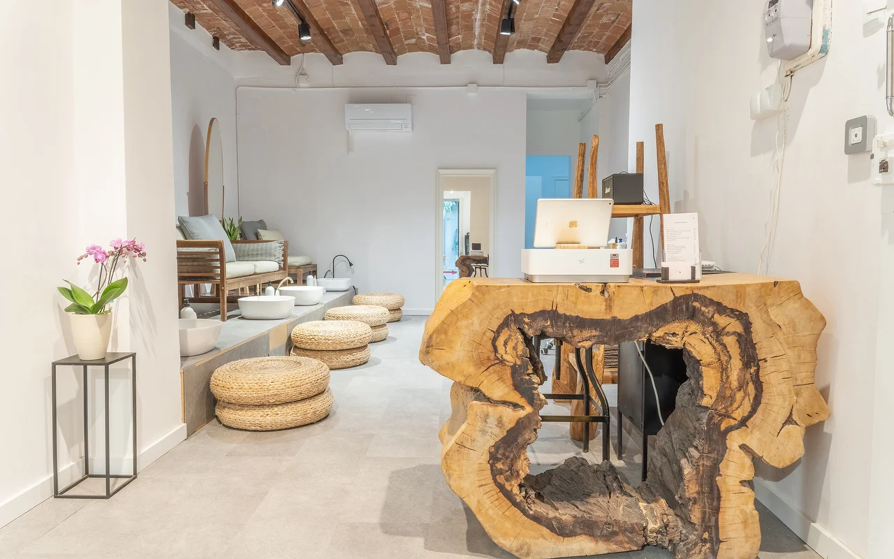 Uno spazio wellness minimalista con un bancone reception ricavato da una grande sezione di tronco d'albero. Sullo sfondo, una postazione pedicure con cuscini da pavimento intrecciati e bacinelle bianche sotto un soffitto rustico con travi a vista.