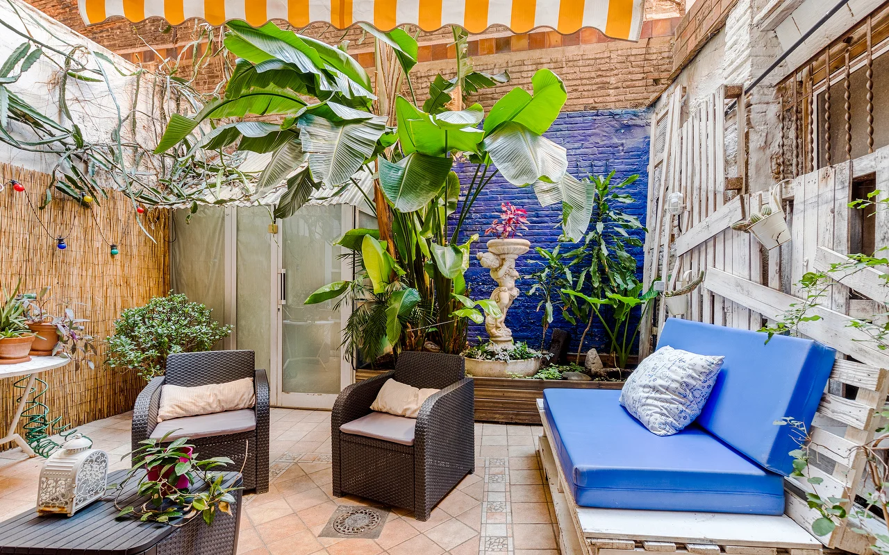 Un eclettico patio esterno o zona d'attesa con una vivace parete blu e rigogliose piante tropicali. Lo spazio comprende poltrone in vimini scuro, una panca con cuscini blu accesi e una tenda a righe gialle e bianche