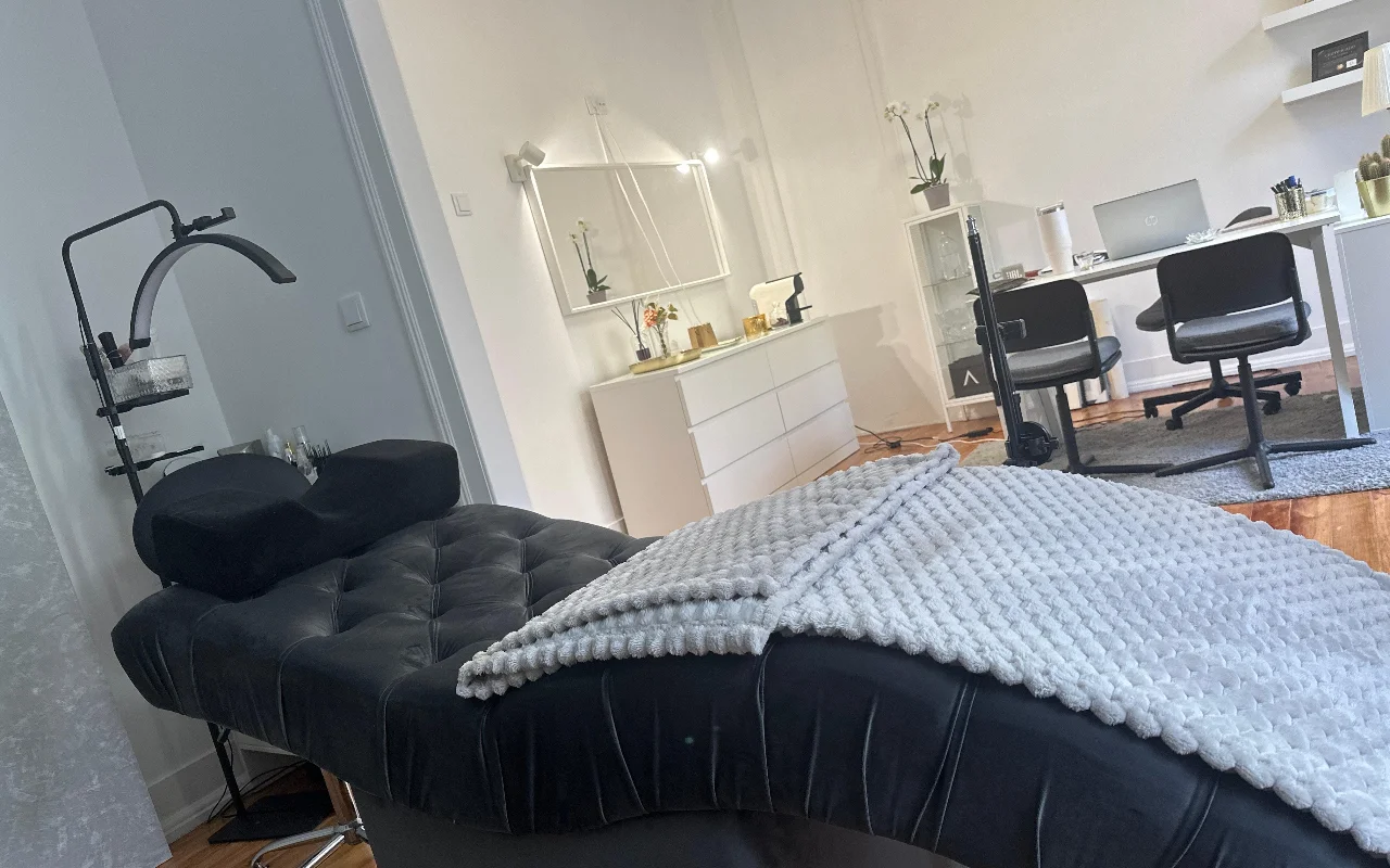 Una moderna stanza per trattamenti ciglia ripresa da un'angolazione bassa. In primo piano, un comodo lettino nero imbottito è drappeggiato con una coperta grigia testurizzata e dotato di un cuscino ergonomico per il collo sotto una ring light circolare. Sullo sfondo, un allestimento minimalista bianco con scrivania, due sedie nere, un laptop, una cassettiera bianca e un grande specchio.