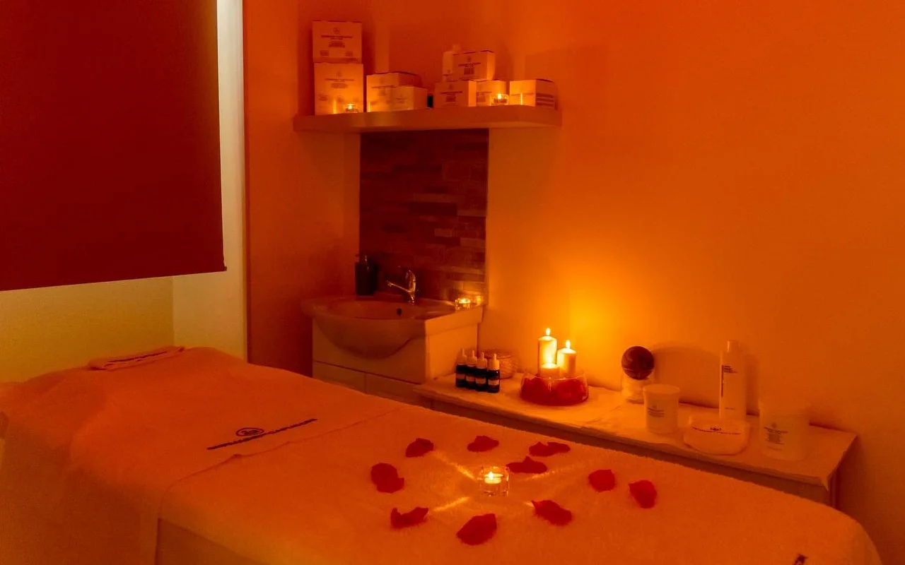 Una sala massaggi con un'illuminazione calda e avvolgente color arancio e candele accese. Petali di rosa rossa sono disposti in un motivo circolare attorno a una candela centrale sul lettino, con prodotti di bellezza ordinati su una mensola sullo sfondo.