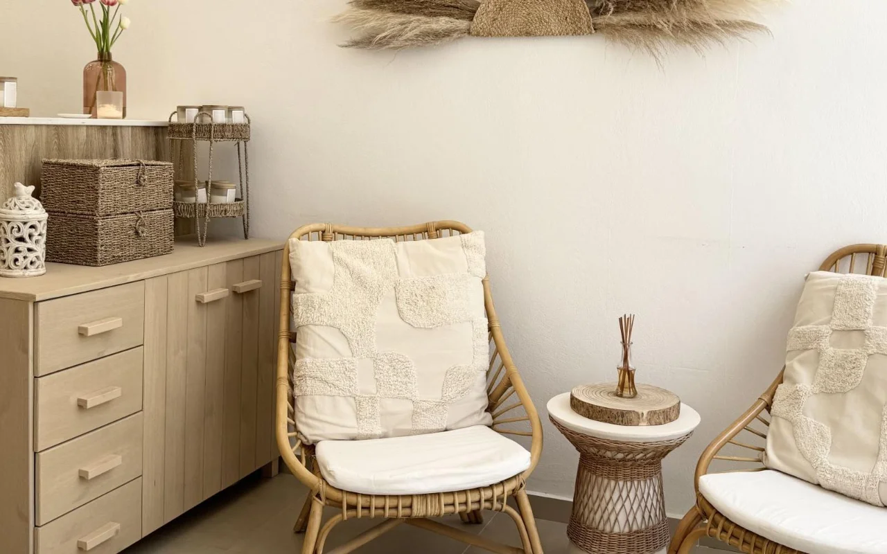 Un'area di attesa rilassante con poltroncine in rattan, cuscini in tessuto crema, un tavolino in vimini con un diffusore a bastoncini, cestini portaoggetti intrecciati e foglie di pampas essiccate sulla parete sopra un mobile in legno chiaro.