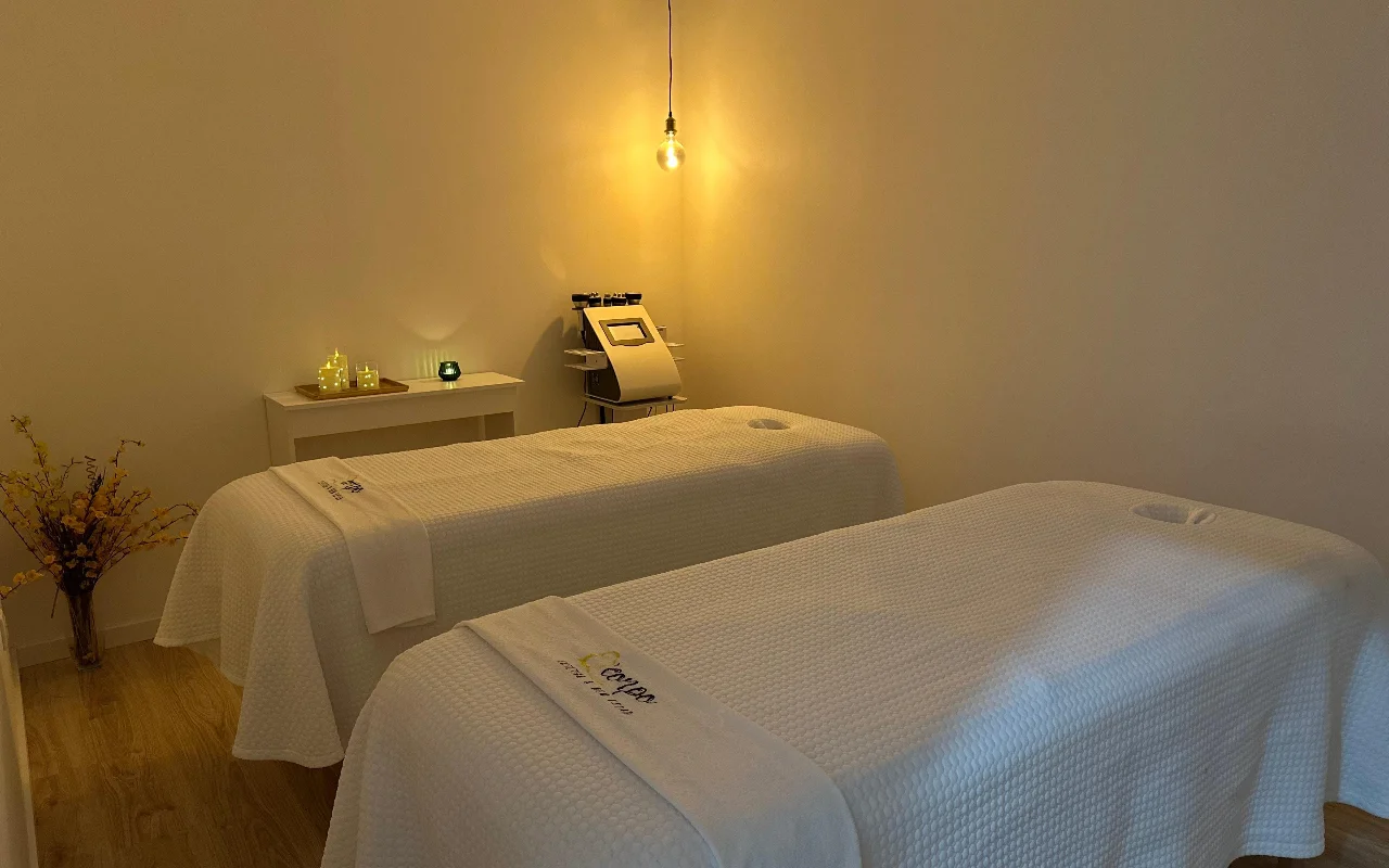 Una tranquilla stanza per massaggi o trattamenti con due lettini affiancati, vestiti con asciugamani e lenzuola bianchi. La stanza è illuminata da una singola lampadina calda a sospensione e piccole candele su un tavolino laterale, creando un'atmosfera serena. Sullo sfondo, un macchinario cosmetico specializzato si trova tra i lettini.