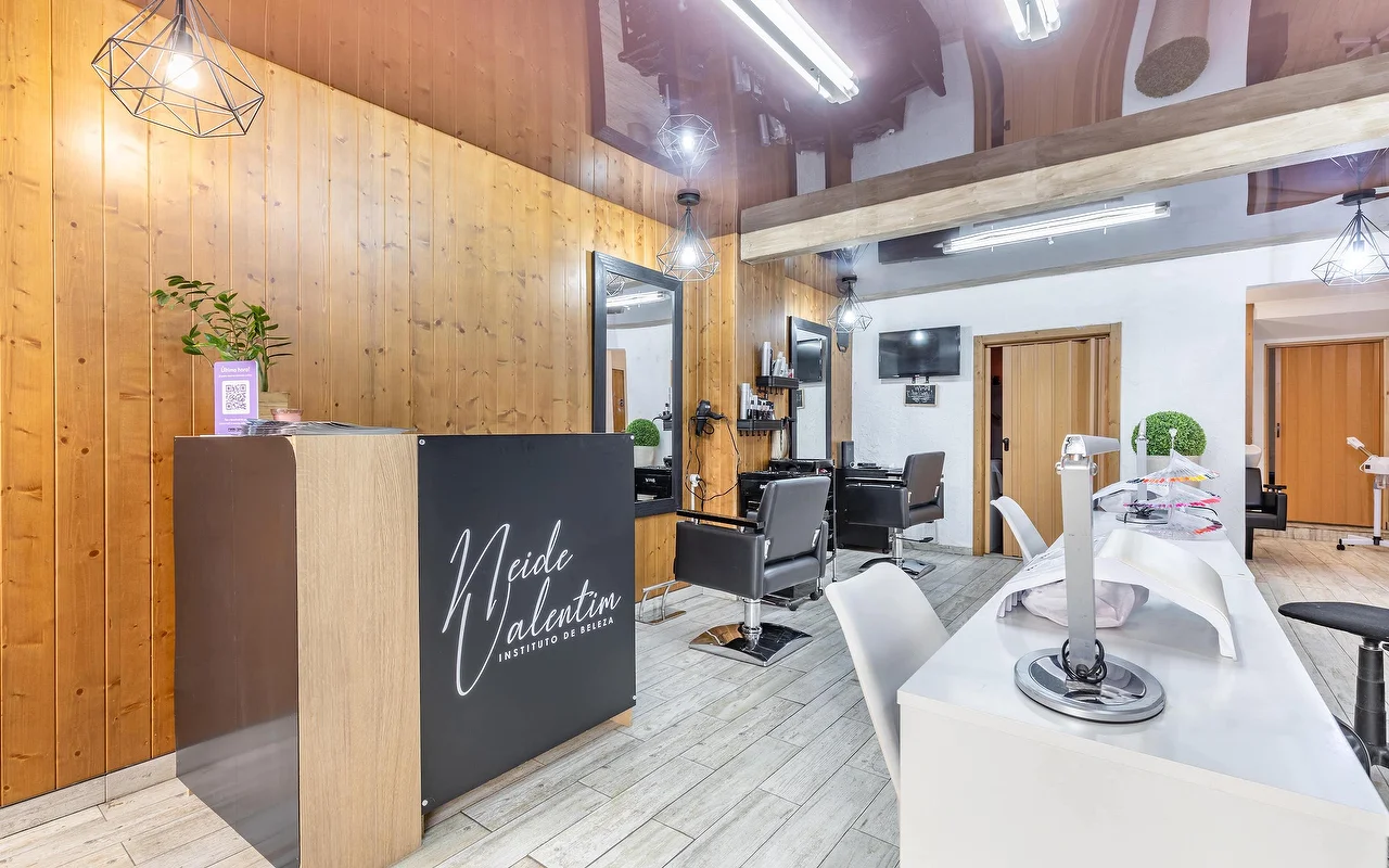 Un moderno salone di bellezza con un bancone reception sulla sinistra brandizzato 'Neide Valentim Instituto de Beleza'. Le pareti sono rifinite con calde boiserie verticali in legno e grandi specchi. Poltrone da parrucchiere nere si trovano davanti agli specchi, mentre una postazione manicure bianca con sedie bianche corre lungo il lato destro del pavimento in piastrelle grigie.