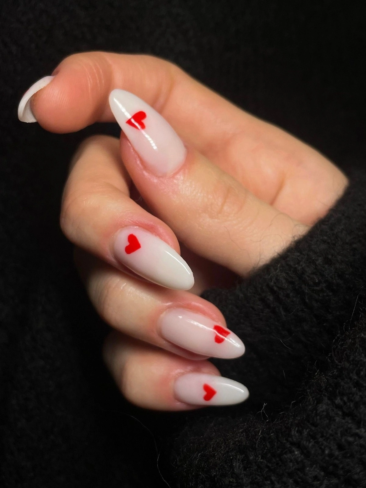 Primo piano della mano di una donna con unghie acriliche bianche a forma di mandorla, decorate con piccoli cuori rossi