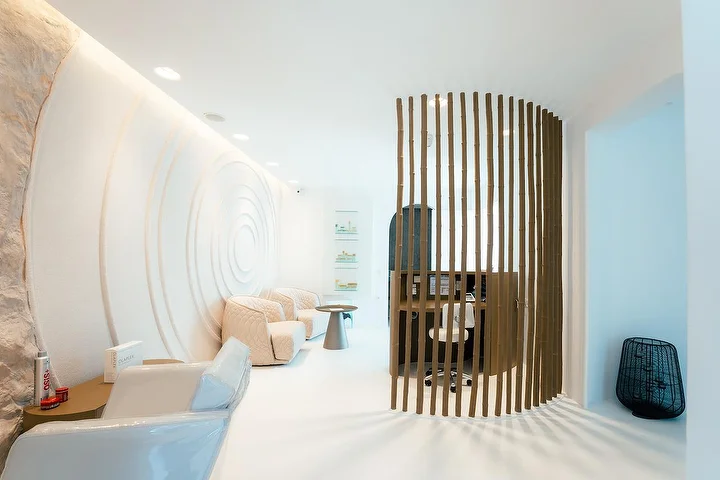 Un interno di spa tranquillo dall'aria di grotta, con pareti bianche curve, una delle quali scolpita in onde concentriche, illuminazione indiretta soffusa, poltrone trapuntate color crema e un paravento curvo a doghe di legno che delimita un angolo trattamenti riservato.