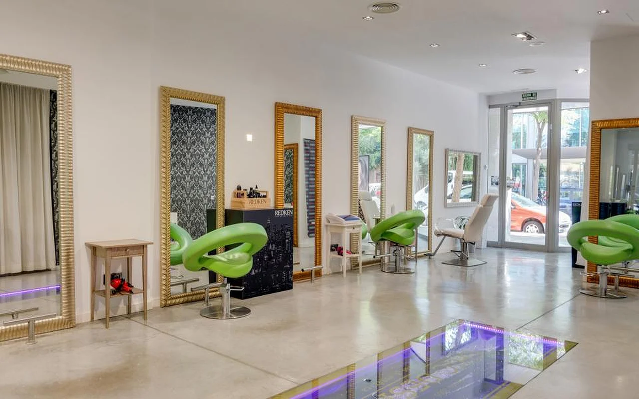 Salone di parrucchiere luminoso e spazioso, con pavimenti in cemento levigato, alti specchi dalla cornice dorata lungo una parete bianca, poltrone da styling sagomate color verde lime e una sezione dal pavimento in vetro illuminata di viola, con la strada visibile dalle vetrate sul fondo.