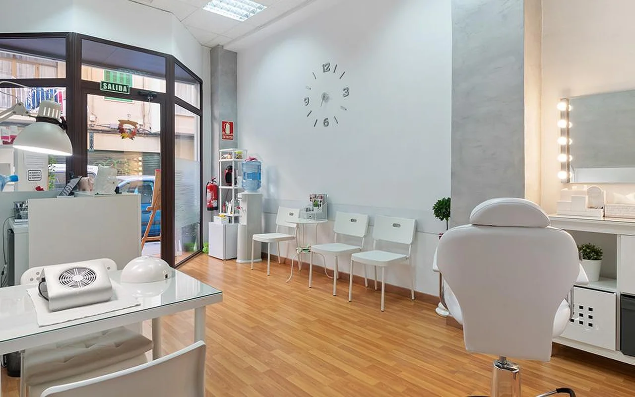 Salone bianco e luminoso con pavimento in legno chiaro, una fila di sedie d'attesa bianche, un orologio da parete minimalista, uno specchio da toeletta illuminato, una poltrona da trattamento reclinabile bianca e una postazione per la manicure vicino alla finestra.