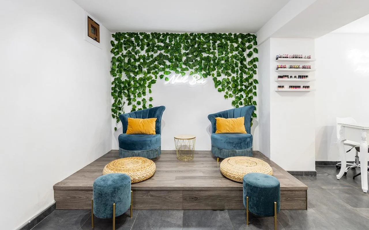 Angolo di un nail bar con una parete di fogliame verde a cascata e una piccola insegna al neon, due poltrone in velluto verde acqua con cuscini color senape su una pedana di legno rialzata, pouf in fibra intrecciata e, sulla destra, mensole di smalti colorati.