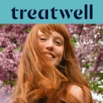 Primo piano ravvicinato di una giovane donna con i capelli rossi e sorridente con la frangetta, con i capelli mossi dal vento davanti a uno sfondo di fiori di lillà rosa. In alto, una fascia azzurra mostra il logo "Treatwell" in blu scuro.