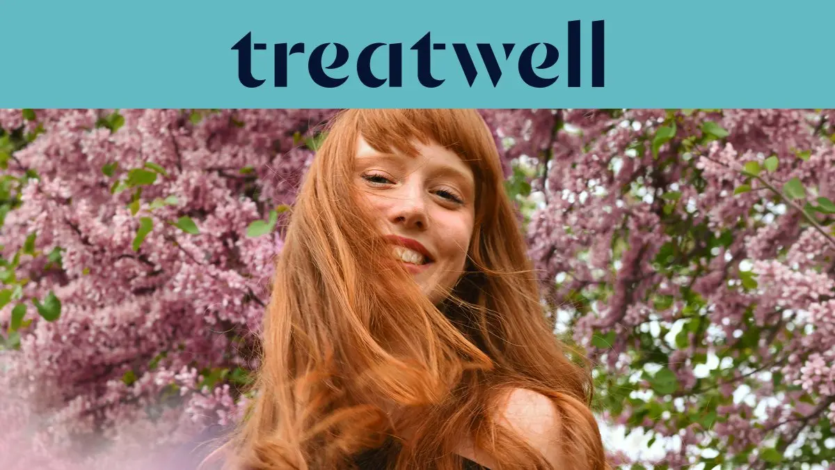 Primo piano ravvicinato di una giovane donna con i capelli rossi e sorridente con la frangetta, con i capelli mossi dal vento davanti a uno sfondo di fiori di lillà rosa. In alto, una fascia azzurra mostra il logo 'Treatwell' in blu scuro.