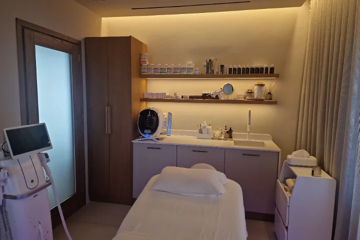 Una cabina trattamenti dalla luce calda, con un lettino viso bianco e asciugamani piegati con cura, scaffalature in legno scuro piene di prodotti per la pelle, uno schermo per l'analisi della pelle e apparecchiature per i trattamenti accanto, clinica ma accogliente.