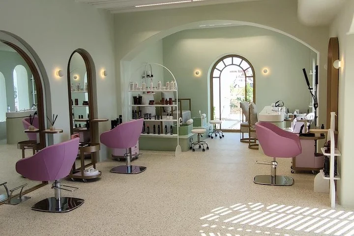 Un salone luminoso verde salvia con pavimento in terrazzo, specchi ad arco con cornice di legno e una finestra ad arco sul giardino, poltrone da styling rosa cipria e mensole aperte piene di prodotti, con la luce del sole che cade sul pavimento.
