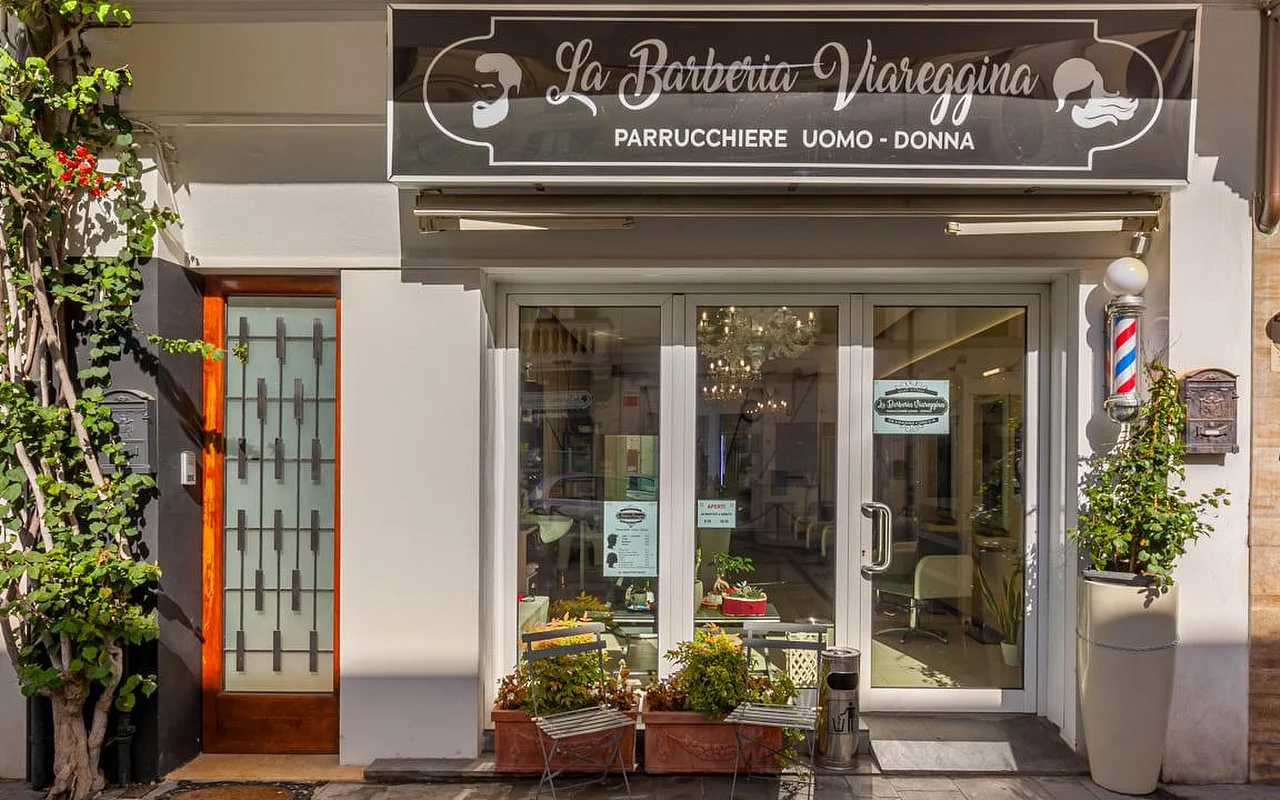 La vetrina de La Barberia Viareggina in una giornata di sole, con la classica insegna rotante da barbiere rossa, bianca e blu a destra delle porte a vetri. Piante in vaso e rampicanti incorniciano l'ingresso, e all'interno, attraverso le vetrine, si intravede un lampadario.