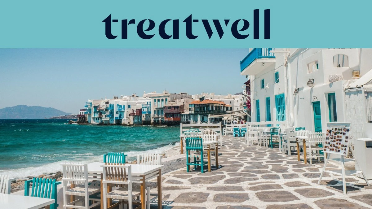 Il lungomare della Piccola Venezia a Mykonos: case imbiancate a calce con balconi di legno colorati a filo dell'acqua, il mar Egeo turchese sullo sfondo e, in primo piano, tavolini da caffè bianchi e verde acqua su una terrazza lastricata in pietra.