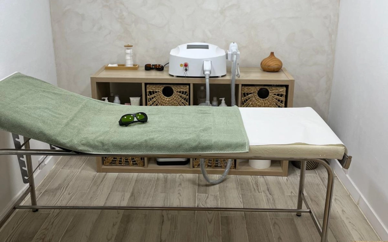 Sala trattamenti dai toni neutri con un lettino dalla struttura in metallo coperto da un telo verde salvia, un paio di occhiali protettivi scuri appoggiati sopra, un macchinario laser su una consolle di legno e cesti portaoggetti in fibra intrecciata sotto.