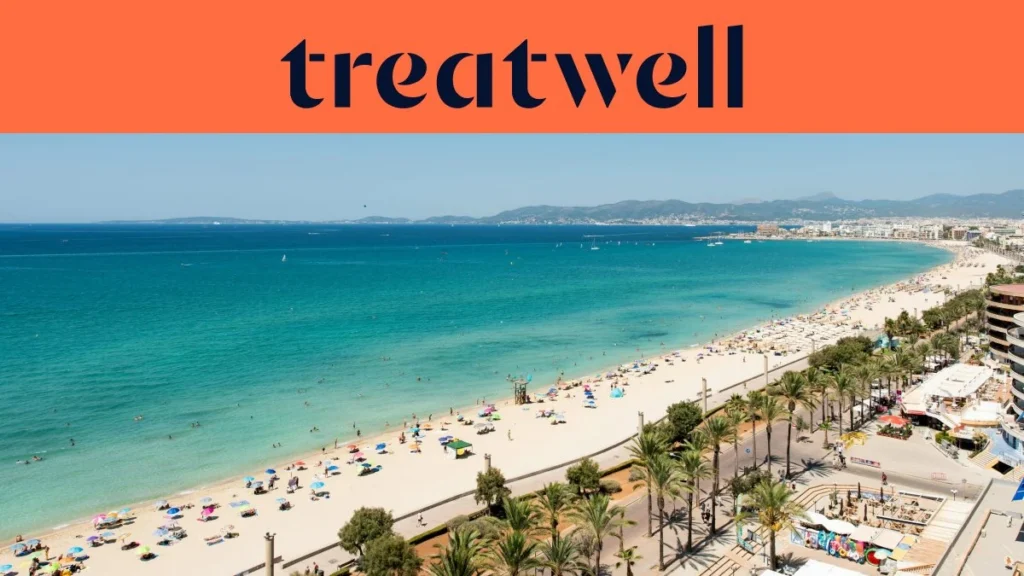 Veduta aerea di Playa de Palma a Maiorca, con una lunga distesa di sabbia dorata, file di ombrelloni, acqua turchese e calma e un lungomare costeggiato di palme che corre lungo la città, con le colline in lontananza sotto un cielo azzurro e limpido.