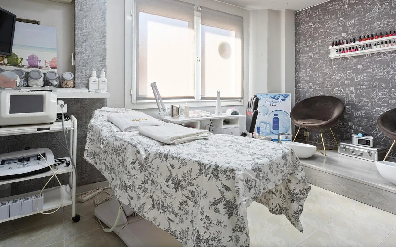 Luminosa sala per trattamenti estetici con un lettino vestito di lino grigio a fiori e asciugamani bianchi piegati, macchinari estetici su carrelli sulla sinistra, una finestra con la tenda a metà e una zona rialzata con una poltrona in velluto marrone e bacinelle per la pedicure davanti a una parete decorata con motivi calligrafici.