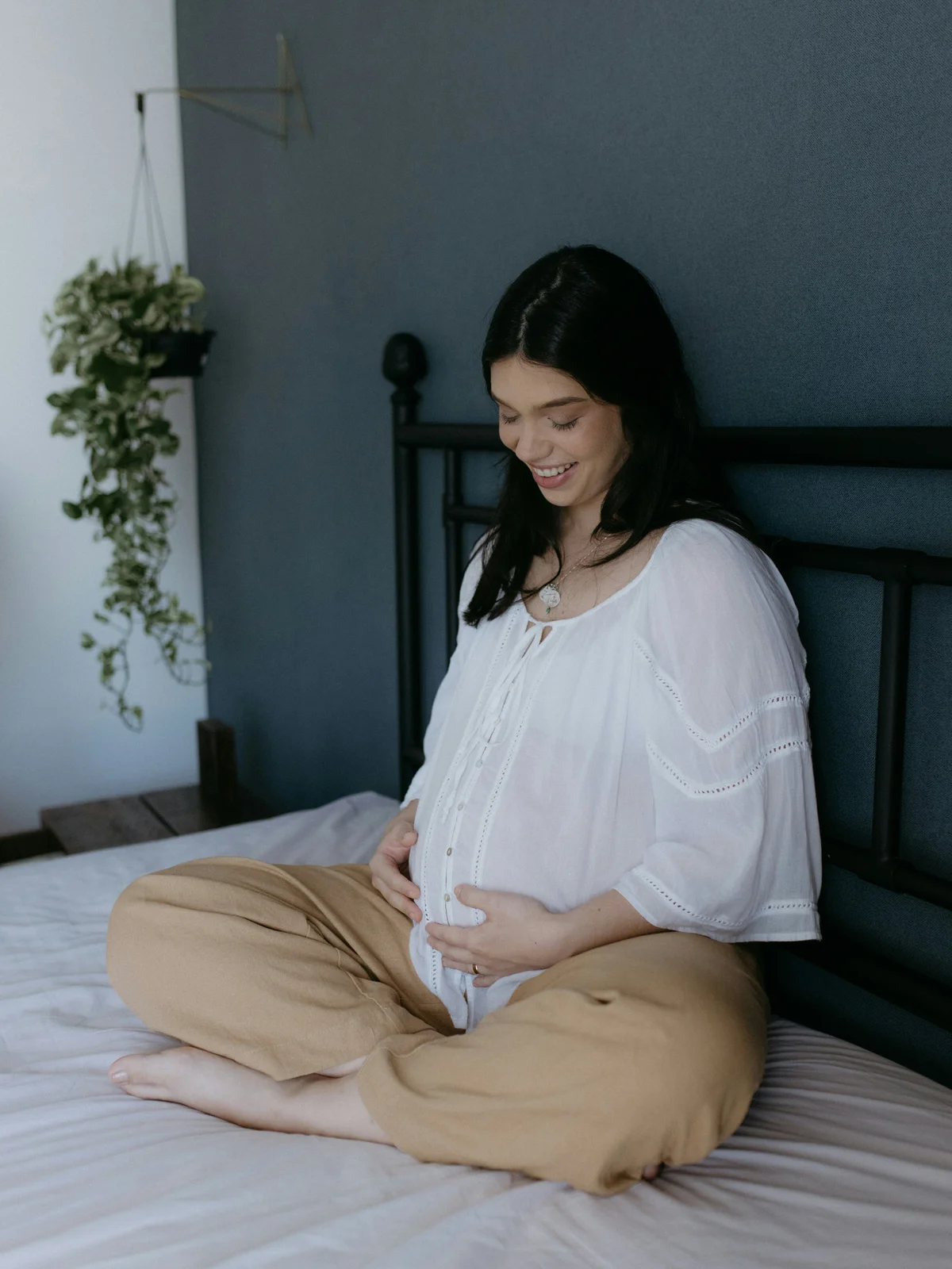 Una donna incinta seduta su un letto a gambe incrociate, con camicia bianca e pantaloni beige, le mani sul pancione e un sorriso rivolto verso il basso