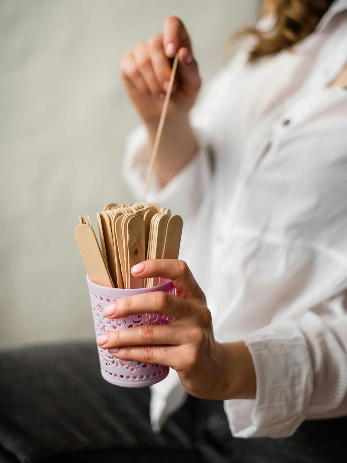 Primo piano di una donna con camicia bianca che tiene con una mano un barattolo rosa pieno di spatole di legno e con l'altra ne estrae una