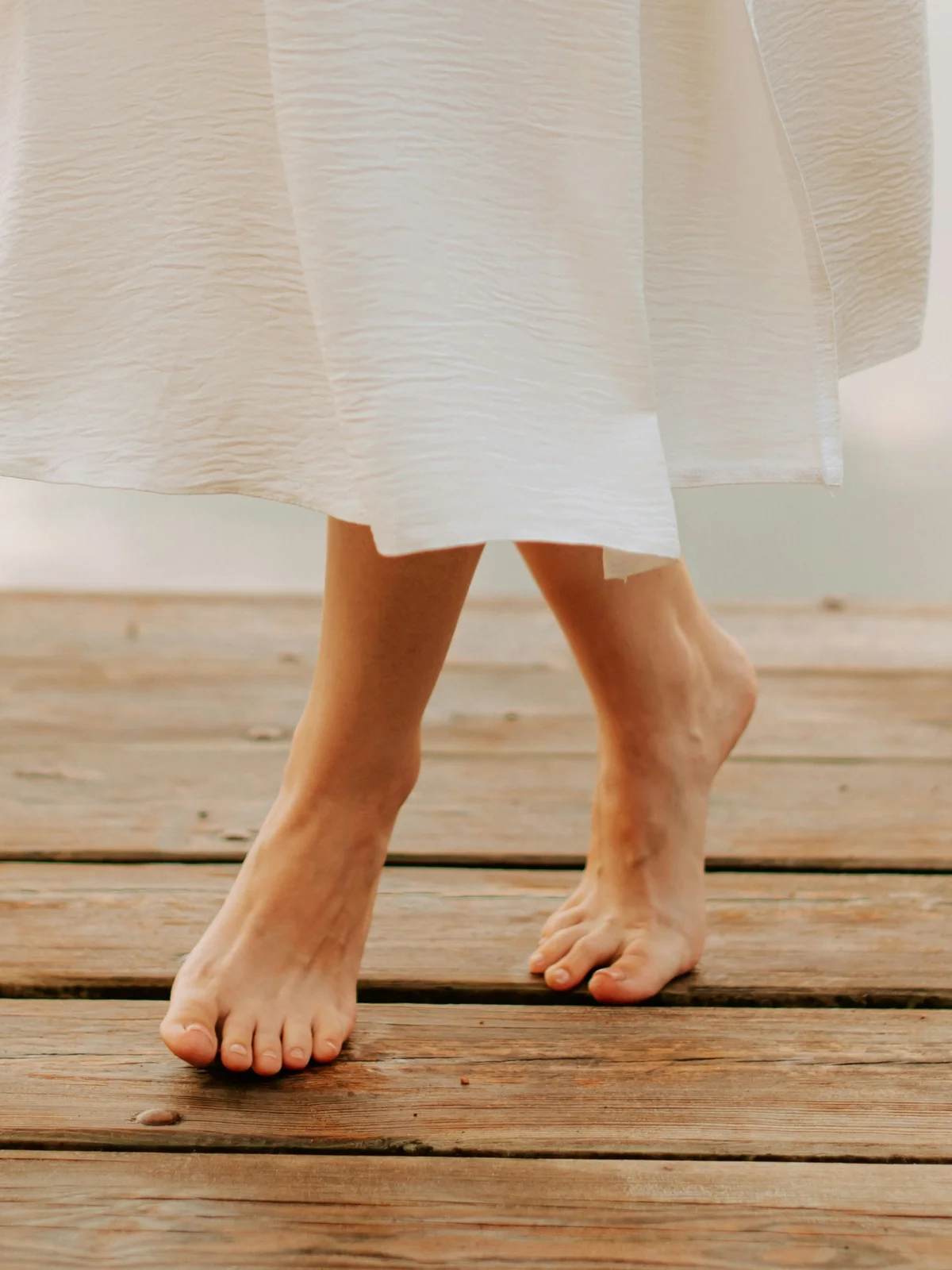Primo piano delle gambe di una donna con piedi nudi e sani e una gonna bianca leggera, mentre fa un passo avanti su una passerella di legno in riva a un lago