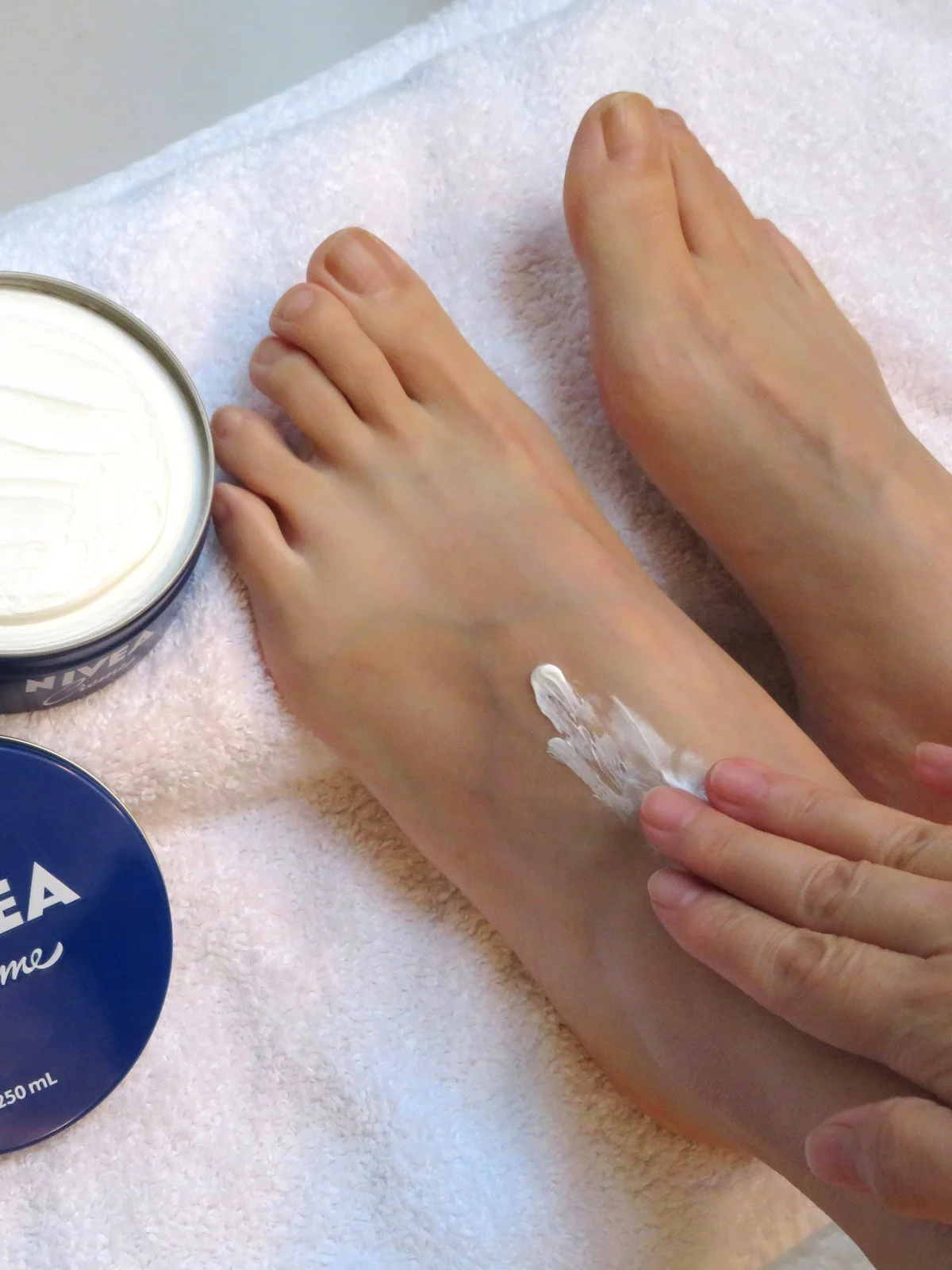 Primo piano dall'alto dei piedi di una donna appoggiati su un lenzuolo bianco, con un vasetto di crema aperto accanto, mentre applica la crema
