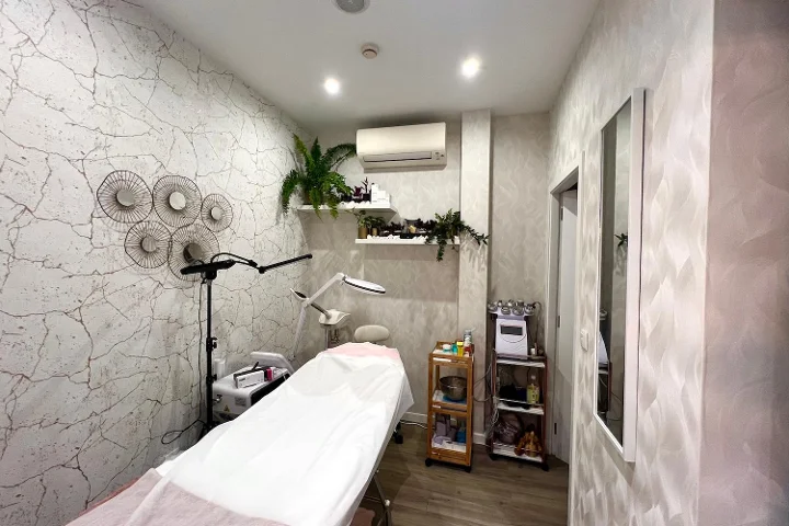 Cabina estetica tranquilla con un lettino trattamenti coperto, una lampada d'ingrandimento e un vaporizzatore, un carrello con i prodotti, pareti testurizzate grigio pallido decorate con dischi metallici e piante ricadenti sopra gli scaffali.