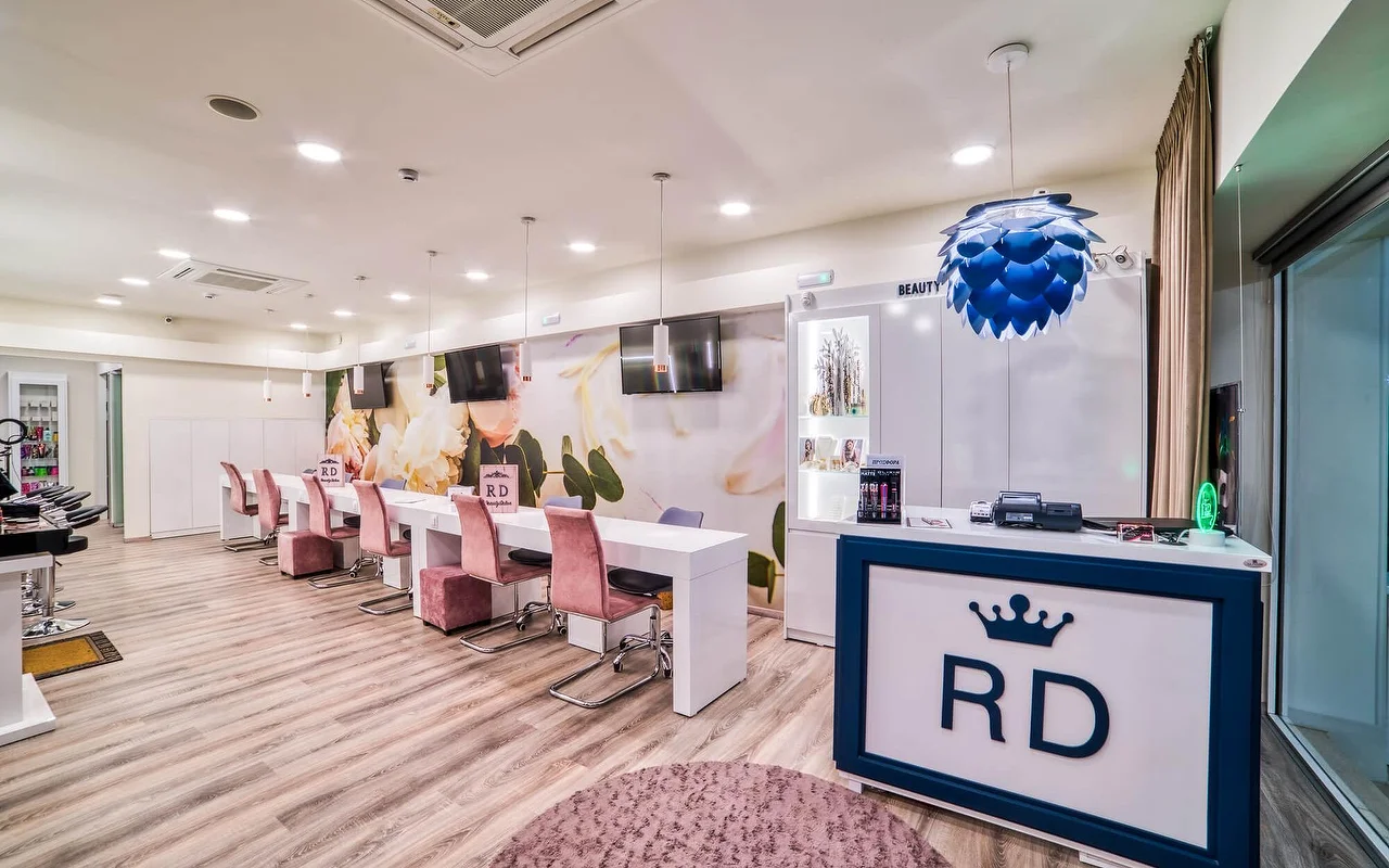 Un salone luminoso e bianco con una lunga fila di tavoli manicure e sedie rosa antico, un delicato murale floreale a parete, pavimento in legno chiaro e un bancone reception con logo a corona 'RD'.