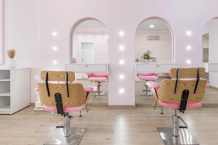 Salone di capelli luminoso in rosa tenue, con poltrone da parrucchiere in legno e tessuto rosa di fronte a due nicchie ad arco illuminate da faretti, mobili bianchi e pavimento effetto legno chiaro.