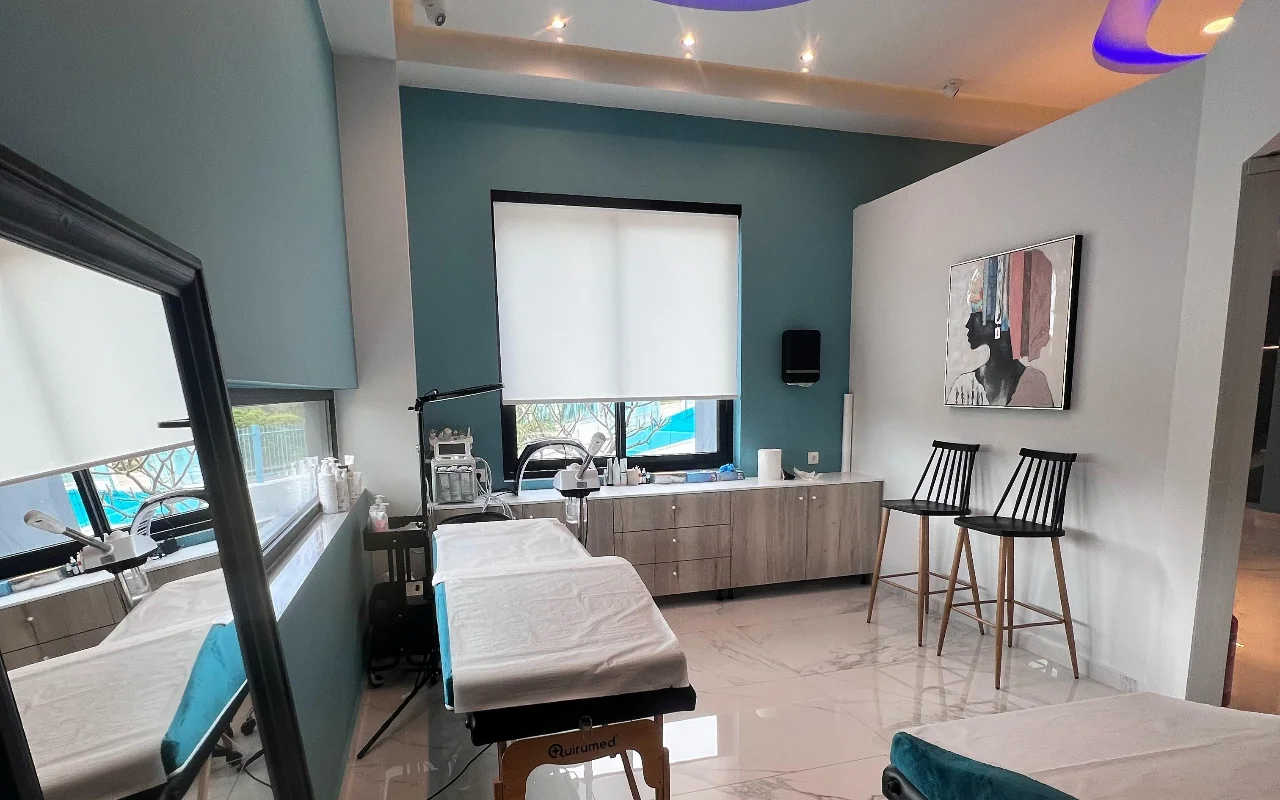 Una sala trattamenti viso con pareti verde acqua, un lettino trattamenti bianco, macchinari skincare, un grande specchio, due sgabelli neri a listelli e un ritratto incorniciato, illuminata da una finestra con tenda a rullo bianca.