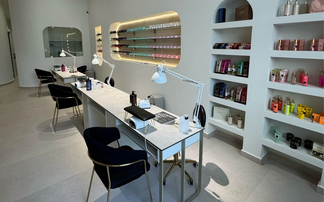 Uno studio unghie bianco con tavoli manicure con piano in marmo e lampade snodabili bianche, sedie in velluto nero su gambe dorate e nicchie a parete ad arco che espongono prodotti skincare e uno scaffale retroilluminato di smalti.