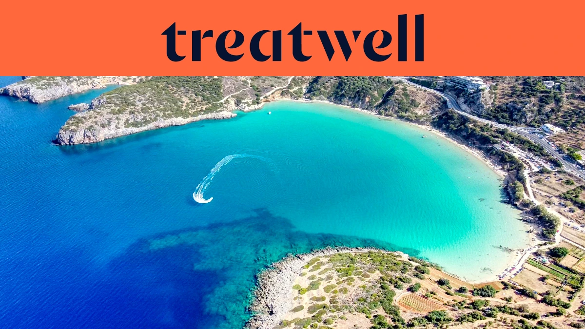 Vista aerea di una baia cretese turchese, con una moto d'acqua che traccia una curva bianca nell'acqua limpida, una mezzaluna di spiaggia sabbiosa e colline aride intorno alla riva, sotto un banner arancione corallo con la scritta treatwell.