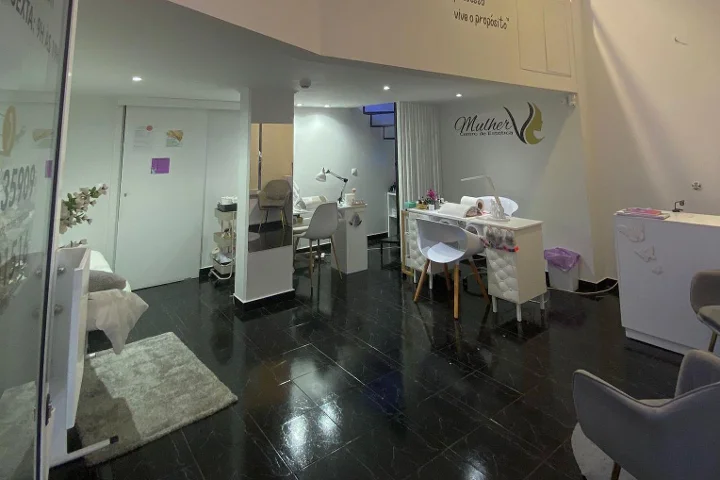 Interno di un centro di bellezza: pavimento di piastrelle scure e lucide, postazione manicure bianca e aree trattamento con luci soffuse, il logo Mulher sulla parete di fondo e un lettino viso visibile dietro una parete di vetro.