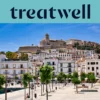 Vista soleggiata su Ibiza città, con edifici imbiancati a calce che salgono fino alla cattedrale e alle antiche mura fortificate di Dalt Vila sulla collina. Una passeggiata del porto alberata attraversa il primo piano sotto un cielo azzurro limpido.