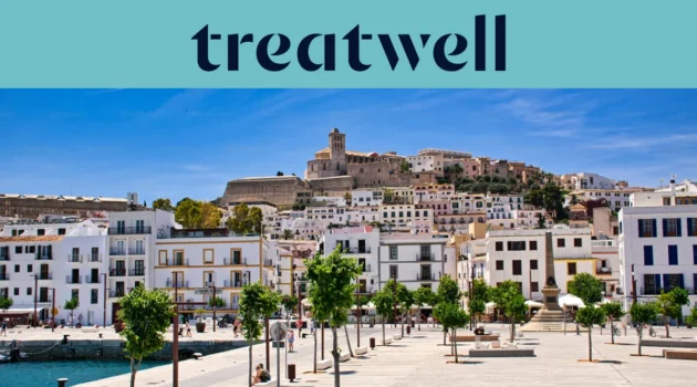 Vista soleggiata su Ibiza città, con edifici imbiancati a calce che salgono fino alla cattedrale e alle antiche mura fortificate di Dalt Vila sulla collina. Una passeggiata del porto alberata attraversa il primo piano sotto un cielo azzurro limpido.