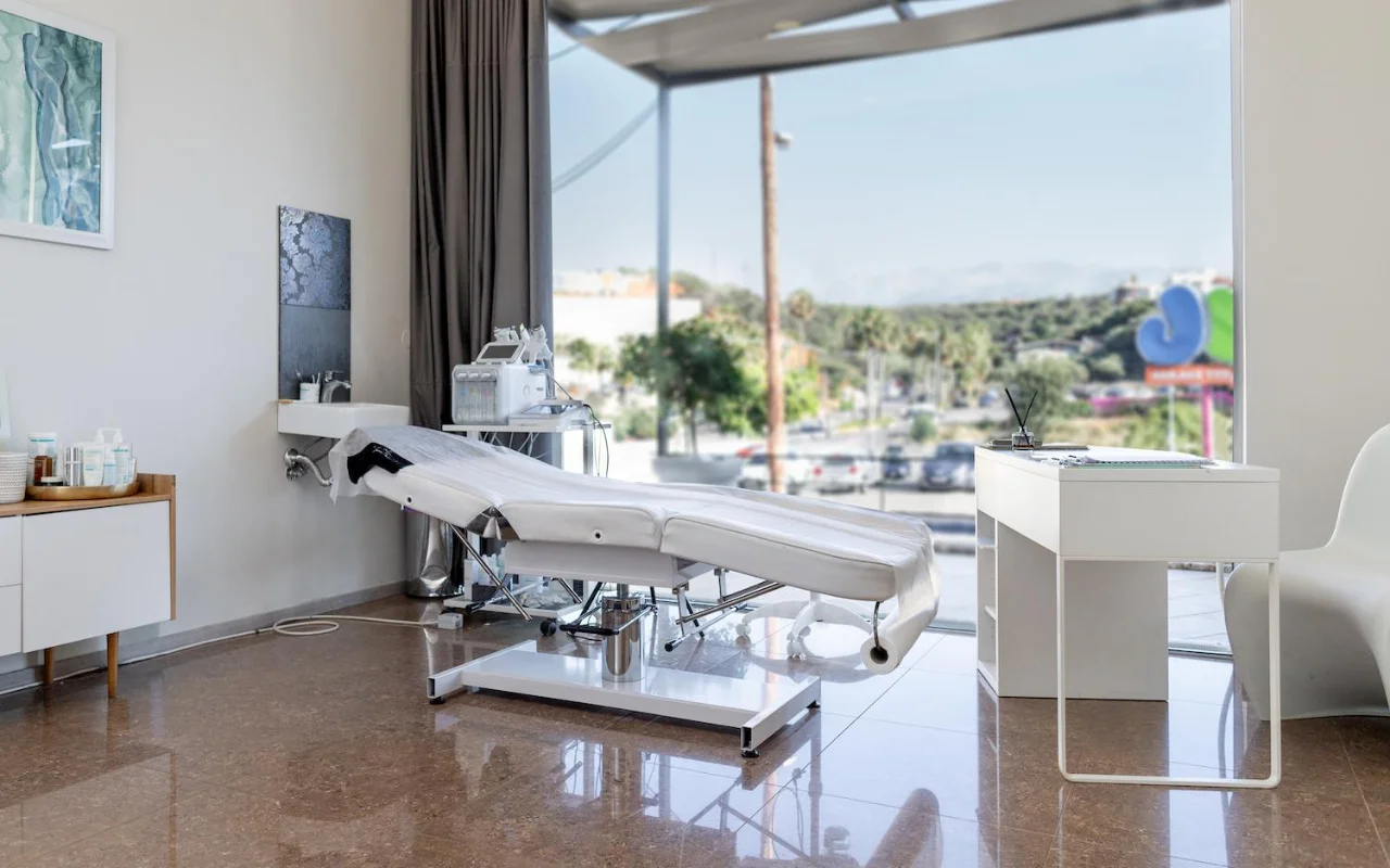 Una sala trattamenti bianca e pulita con un lettino estetico reclinabile, apparecchiature skincare su un carrello, opere d'arte incorniciate e una grande finestra affacciata su alberi, una strada e colline in lontananza.