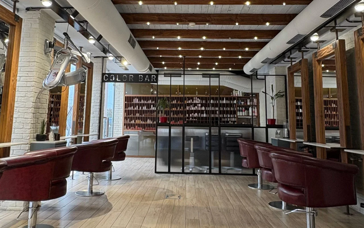 Un salone di parrucchiere con travi a vista in legno e muratura imbiancata, poltrone da hairstyling in pelle bordeaux e una sala colore con pareti in vetro rivestita di scaffali di prodotti dietro un'insegna 'Color Bar'.